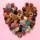 Photograph showing a variety of knitted baked goods, including tarts, biscuits, macarons and gingerbread men arranged in the shape of a heart on a pink background