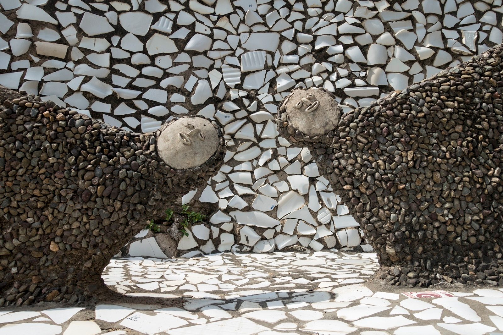 Two somewhat abstract human figures made from discarded pottery fragments lean into the photo. Both the floor and background wall are inlaid with large pieces of discarded white pottery.