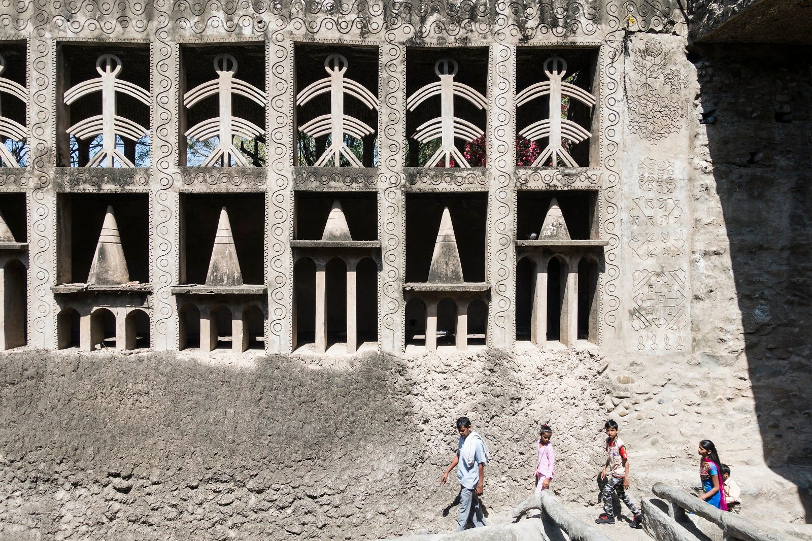 Three men stand beneath a huge concrete wall with recesses filled with abstract sculptures.