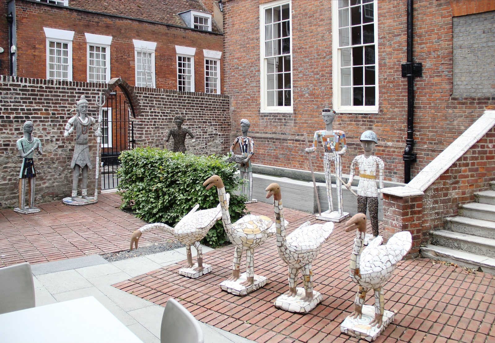 A set of four swan sculptures stand in a row in front a row os sculptures of human figures. All the sculptures are made from concrete and discarded pottery fragments