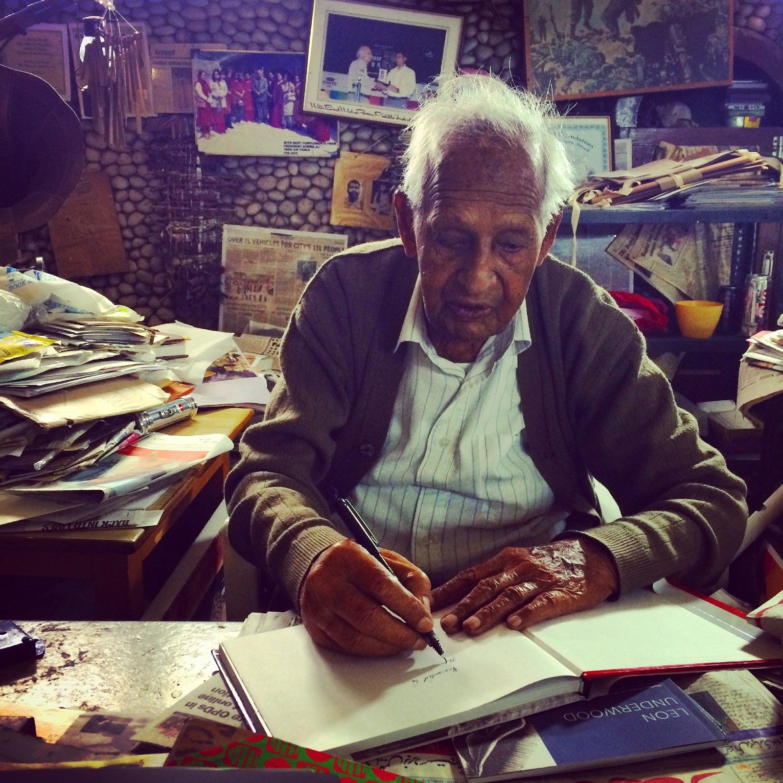 An elderly Indian man draws ina sketchbook surrounded by piles of paper, photographs and other ephemera