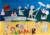 A collaged beach scene, depicting people sunbathing, enjoying an ice cream, building sandcastles and reading on the sand. On the water is a life buoy, the QE2 cruiseliner, two people paddleboarding and a swimmer with both hands in the air and a speech bubble saying 