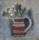 A painting showing a flattened image of a jug with red, white and black stripes containing a bunch of wild flowers.