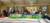 Photograph of a group of around 20 people stood behind a long artwork showing a green landscape and blue sky