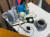 Painting of a white table with a grey cup and saucer with coffee in the cup, a plate with a knife lying across it and a small piece of cake and at the back a grey elephant plush toy and next to it a rabbit plush toy. Next to them are blue and red leaflets in a stand.