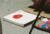 Photograph of a pad of paper with a painting of a red tomato on top with a tray of platic balls in many colours next to it on the floor