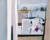 Painting of a view from a window with a jug on the window sill and in the back ground you can see sailing ships on the sea with mountains behind them