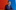 Photograph of a man leaning his elbows on the edge of a waist high wall. He wears a blue suit with white shirt and a red tie. In the background is a wall of many different coloured triangles.