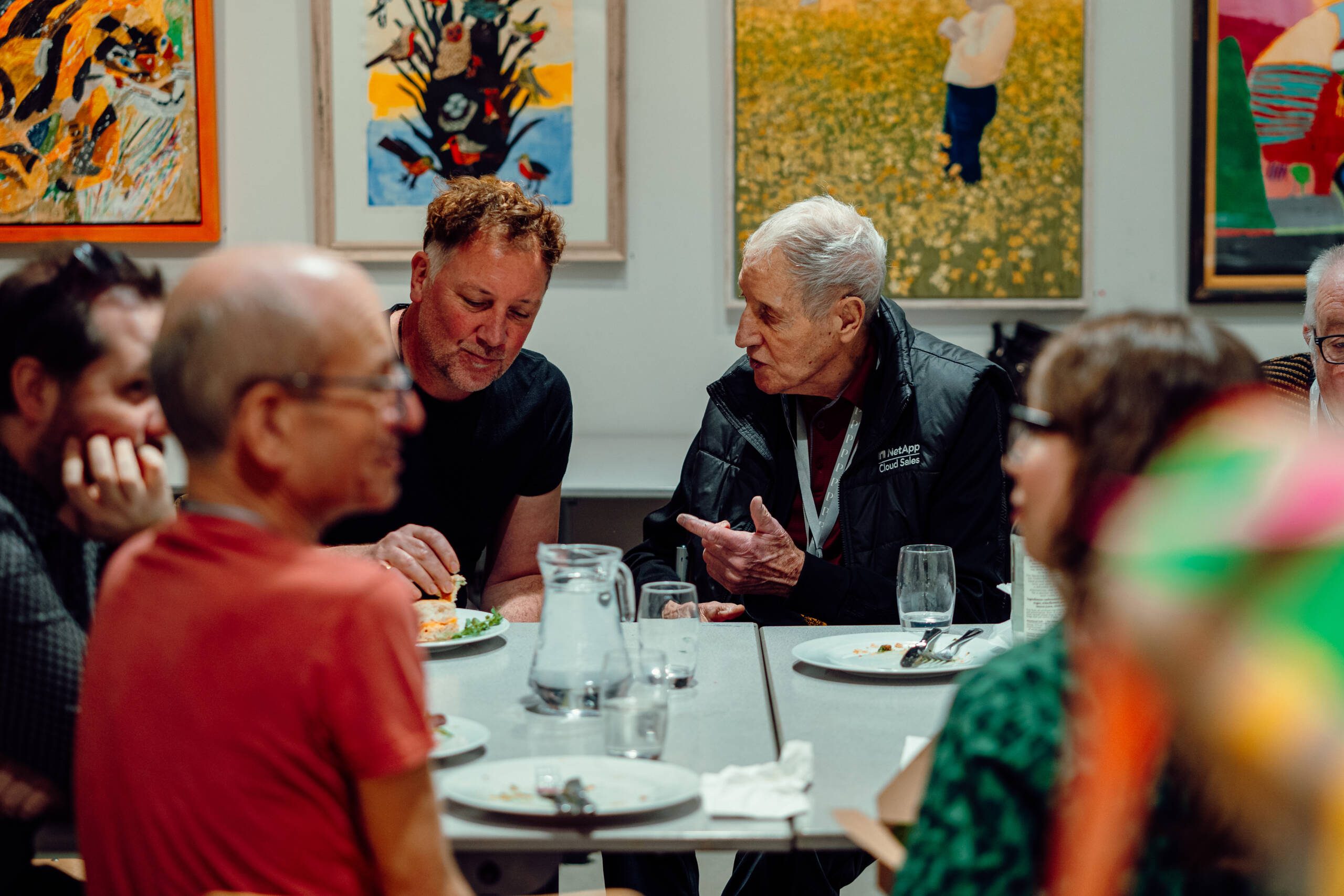 Photograph of a group of people sat at a table chatting with artworks on the wall behind them